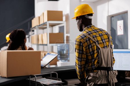 Photo for Courier checking orders list and receiving parcel in retail storehouse. African american warehouse managers preparing package delivery invoice at reception desk in storage room - Royalty Free Image