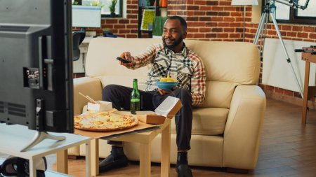Photo for African american adult drinking beer and eating chips, enjoying comedy tv show in living room. Modern man having fun with movie on television, serving takeaway fast food at home. - Royalty Free Image