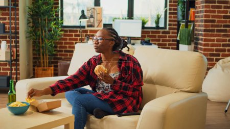 Téléchargez les photos : African american person taking bite of burger on couch, eating fast food from delivery a home. Young adult enjoying takeaway meal with fries and hamburger, watch movie on television. - en image libre de droit
