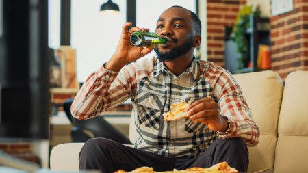 Photo for Cheerful young guy drinking beer and eating pizza, watching film on channel program at home. Happy man enjoying dinner with fast food takeout meal and alcohol, watch movie. Handheld shot. - Royalty Free Image