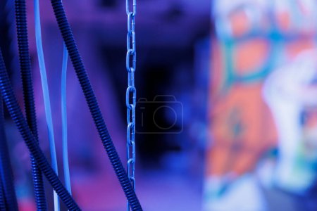 Photo for Chains hanging out of walls in deserted messy space, rusty neglected building. Empty abandoned place filled with bright purple lights and artwork, glowing under fluorescent light. Close up. - Royalty Free Image