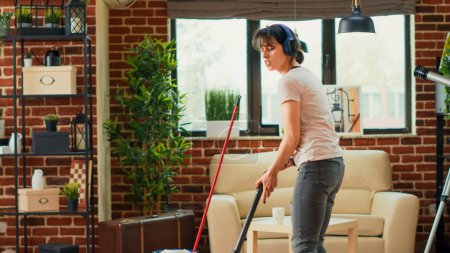 Téléchargez les photos : Female person listening to music on headset and vacuuming floors, having fun with vacuum cleaner. Young modern woman feeling cheerful while she does spring cleaning household. - en image libre de droit