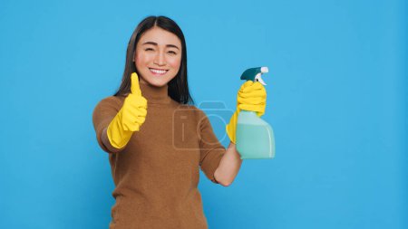Foto de Smiling positive housekeeper holding chemical detergent spray doing approval gesture in studio. Maid is always professional in her cleaning, making sure to pay attention to even the smallest details. - Imagen libre de derechos