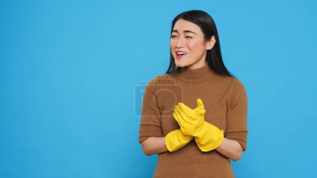 Téléchargez les photos : Souriant asiatique femme de ménage applaudissant mains devant la caméra après avoir terminé de faire le nettoyage de la maison, debout en studio sur fond bleu. Housekepper était très fière de ses tâches ménagères - en image libre de droit