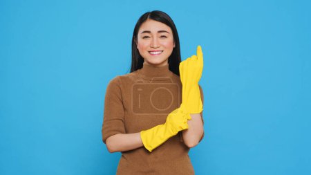Photo for Asian maid smiling at camera while putting rubber glove after finishing house cleaning, standing in studio. Housekepper was skilled in the art of housekeeping, ensuring every surface was clean - Royalty Free Image