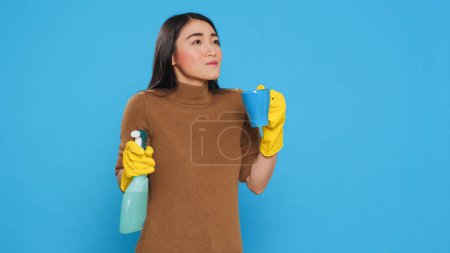 Photo for Happy housekepper enjoying cup of coffee while cleaning house with chemical spray bottle, standing in studio over blue background. Maid using protective equipment and sanitary practices - Royalty Free Image