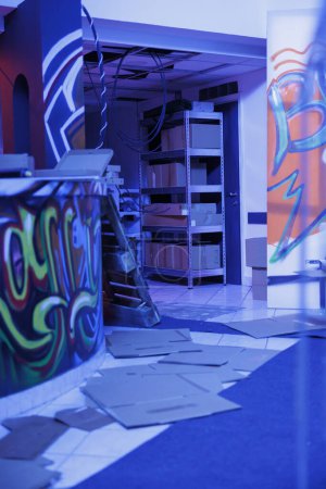 Photo for Urban graffiti artwork glowing under neon lights in deserted messy warehouse, empty building filled with spray paint and garbage, fluorescent bright blue light. Abandoned damaged space. - Royalty Free Image