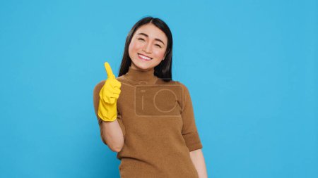 Téléchargez les photos : Smiling maid wearing yellow rubber gloves while doing ok gesture after finishing to clean client house, posing in studio. Cheerful housekeeper is responsible for providing cleaning services - en image libre de droit