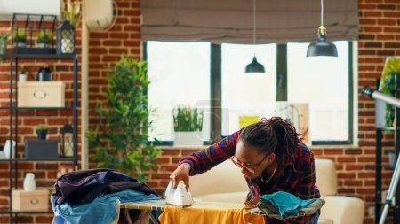 Téléchargez les photos : African american woman using steam iron and ironing board, smoothing out creases on clothes. Female person doing household chores for spring cleaning, using laundered clothing. - en image libre de droit