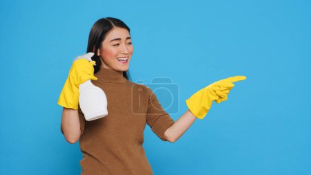 Téléchargez les photos : Cleaning woman pointing and showing cleaning product or isolated text, using spray bottle filled with sanitary solution to disinfect surfaces and prevent the spread of germs. Housecleaning concept - en image libre de droit