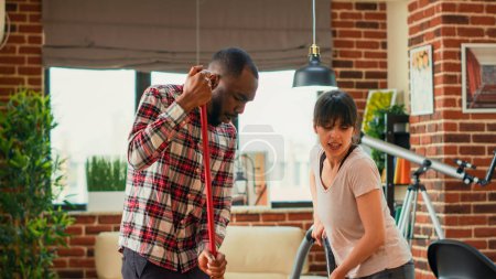 Photo for Diverse people showing dance moves and enjoying spring cleaning, mopping and vacuuming floors in apartment. Happy life partners having fun with mop and vacuum cleaner. Tripod shot. - Royalty Free Image