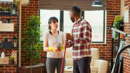 Photo for Diverse couple cleaning apartment with tools and appliances, man mopping wooden floors and woman sweeping dust off of furniture. Life partners using mop and rags to clean house. Handheld shot. - Royalty Free Image