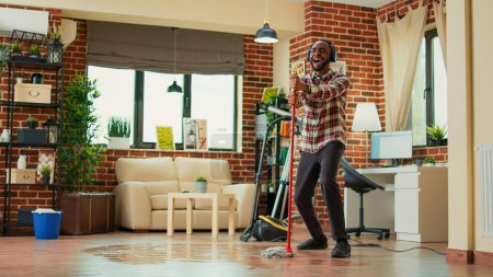 Téléchargez les photos : Young male adult having fun with mop and music at home, showing dance moves and singing in living room. Happy person mopping wooden floors and doing spring cleaning, housework. - en image libre de droit