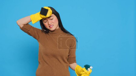 Téléchargez les photos : Overwhelmed maid standing in studio over blue background doing intensive cleaning using chemical detergent spray and sponge. Tired housekepper is dedicated to maintaining the cleanliness - en image libre de droit