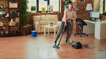 Téléchargez les photos : Happy woman listening to music on headphones and vacuuming floors at home, doing spring cleaning work in living room. Young adult singing and doing dance moves while she uses vacuum, having fun. - en image libre de droit
