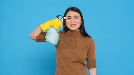 Photo for Upset angry maid putting detergent spray in front of temple while pretending to shoot herself after finishing cleaning customer house. Cleaner was wearing gloves while doing the housework - Royalty Free Image