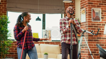 Photo for Funny people showing dance moves and tidying up apartment, mopping and vacuuming floors while they listen to music. Young cheerful couple using mop and vacuum cleaner. Handheld shot. - Royalty Free Image