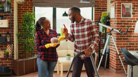 Téléchargez les photos : Young life partners doing household chores and cleaning apartment with vacuum cleaner. African american woman sweeping dust off of furniture with rags and washing solution, furniture polish. - en image libre de droit