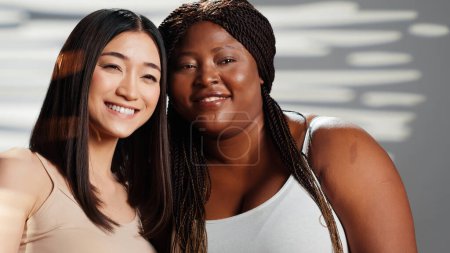 Téléchargez les photos : Interracial girls embracing and expressing self confidence, doing skincare and advertisement in studio. Happy women posing and showing self acceptance, body positivity femininity concept. - en image libre de droit