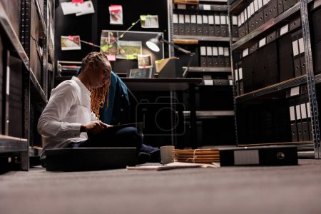 Photo for Lawyer preparing for case and reading detective report while sitting on floor. African american investigator analyzing csi expertise and evidence while working overtime at night - Royalty Free Image