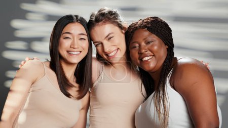 Diverse schöne Mädchen posieren für Körpervertrauensanzeige im Studio, eine Gruppe von Freunden wirbt für Hautpflegeprodukte. Positiv selbstbewusste Frauen, die vor der Kamera lachen, unterschiedliche Hautfarben und Körperformen.