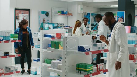 Foto de Pharmacy employee showing medicaments to woman, recommending healthcare products and pills. Young customer talking to pharmacist about medical treatment, pharmaceutics. Tripod shot. - Imagen libre de derechos