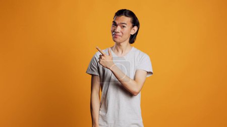 Foto de Asian person pointing sideways with index fingers, looking left or right sides and being confident. Young male model indicating directions aside in studio, posing with confidence on camera. - Imagen libre de derechos