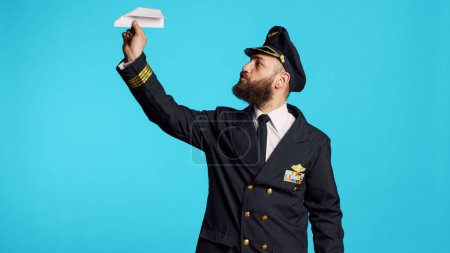Téléchargez les photos : Young man working as pilot building paper plane, having fun with origami in studio. Male captain in aviation uniform playing with folded airplane, having professional occupation as airliner. - en image libre de droit