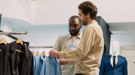 Photo for Shopping center worker helping client with trendy outfits to buy modern clothes in clothing store. Young shopper talking to employee and examining formal wear on hangers. Handheld shot. - Royalty Free Image
