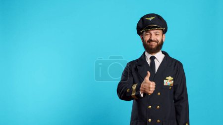Photo for Aviation male pilot giving thumbs up on camera, expressing good sign and approval. Aircrew captain with flying uniform showing agreement and accept, doing okay symbol in studio. - Royalty Free Image