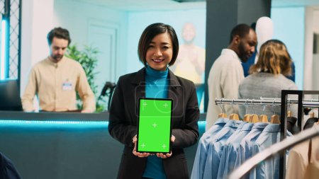 Photo for Asian store assistant using greenscreen display on tablet, holding isolated mockup display in clothing store. Cheerful boutique worker showing chroma key copyspace template in retail market. - Royalty Free Image