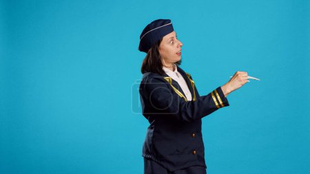 Téléchargez les photos : Caucasian woman playing with plane toy, being dressed as stewardess and showing miniature fake airplane in studio. Person working as air hostess holding artificial mini aircraft over blue background. - en image libre de droit