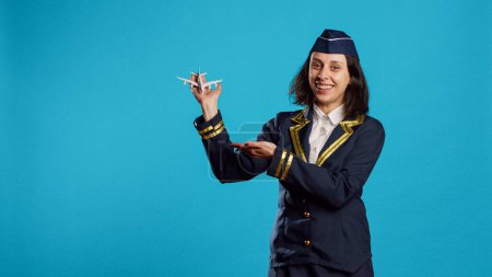 Photo for Professional air hostess presenting miniature toy plane, feeling happy about flying occupation. Young woman working as stewardess in uniform showing small artificial airplane on camera. - Royalty Free Image