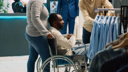 Photo for Young adult with impairment visiting clothing boutique, looking to buy modern casual wear in shopping mall. Male client wheelchair user examining fashionable clothes. Tripod shot. - Royalty Free Image