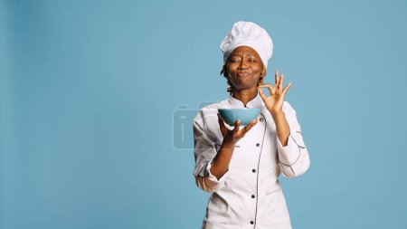 Cheerful gourmet chef smelling food aroma from bowl on camera, enjoying professional meal cooked. Professional female cook savoring tasty food, flavor and scent in studio. Catering service.