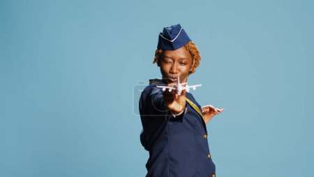 Photo for Professional airliner showing fake miniature plane, playing with artificial mini airplane toy on camera. Confident stewaress having fun with small imitation flying aircraft on blue backdrop. - Royalty Free Image