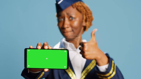Photo for Female aircrew member pointing at greenscreen display over blue background on camera. Young stewardess showing isolated copyspace and chroma key template dressed in aviation uniform - Royalty Free Image