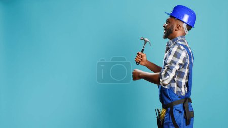 Foto de Carpintero afroamericano golpeando paredes con martillo, utilizando un instrumento manual para trabajos de renovación y reconstrucción. Constructor masculino en overoles sosteniendo herramienta de salmonete jackhammer. - Imagen libre de derechos