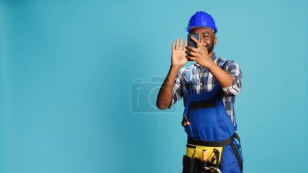 Photo for Cheerful builder talking on video call meeting in studio, using online videoconference chat to talk about new building contract. Young handyman in overalls using remote teleconference. - Royalty Free Image