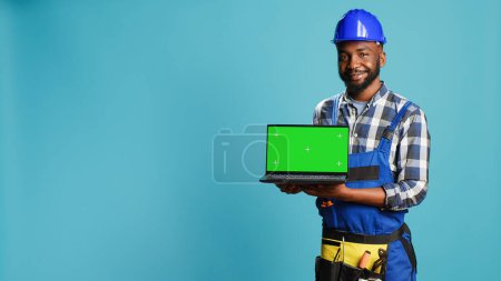 Photo for Smiling craftsman showing laptop with green screen on camera, advertising modern isolated display on pc. Young builder contractor holding computer with blank chroma key copyspace screen. - Royalty Free Image