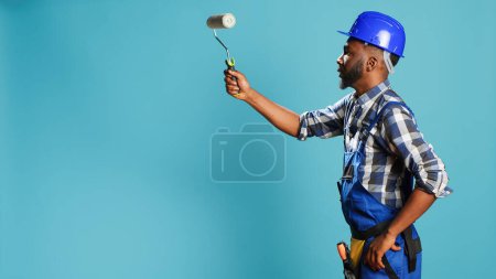 Photo for Industrial worker in uniform painting walls with roller, using renovation instrument over blue backdrop. Professional craftsman with hardhat working with rolling paint brush to change color. - Royalty Free Image