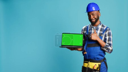 Photo for Happy mechanic pointing at laptop with greenscreen on camera, advertising isolated template display on pc. Young construction worker showing computer with chroma key mockup copyspace. - Royalty Free Image