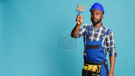 Photo for African american man painting walls with brush in studio, using renovating tools to change color. Professional builder working with paintbrush on carpentry equipment over blue backdrop. - Royalty Free Image