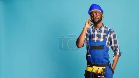 Photo for African american carpenter talking on phone call in studio, planning new construction project on camera. Male builder in overalls using smartphone line to chat remotely, building contract. - Royalty Free Image