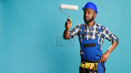Photo for Repairman in overalls painting walls with roller brush, using renovation equipment on camera. Confident carpenter with helmet working with rolling paint brush to change color in studio. - Royalty Free Image
