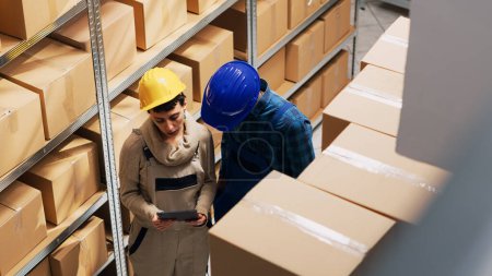 Photo for Diverse people analyzing stock on digital tablet in depot, planning order shipment with merchandise in cardboard boxes. Team of man and woman checking products placed on warehouse racks. - Royalty Free Image