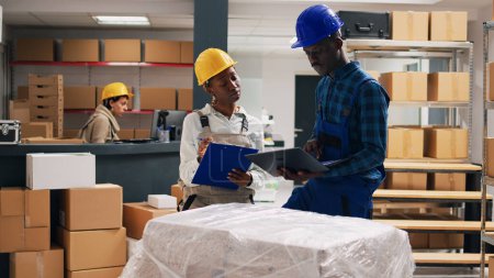 Photo for African american people in overalls analyzing products placed on storage room shelves, cargo shipping in cardboard boxes. Man and woman working with industrial goods, depot space. - Royalty Free Image