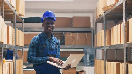 Photo for Warehouse employee counting goods boxes on racks, checking stock list on laptop before products distribution. Young adult in overalls using computer to work on inventory and logistics. - Royalty Free Image