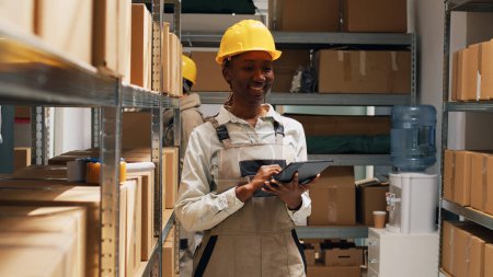 Photo for African american woman in overalls working with tablet, using device to plan stock logistics for warehouse merchandise. Stockroom employee checking industrial cargo in cardboard boxes. - Royalty Free Image