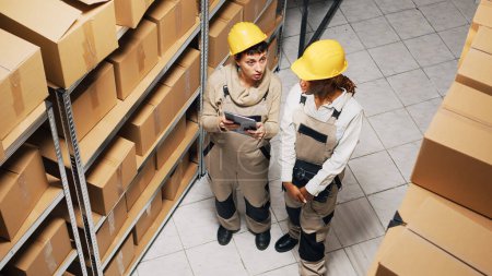Photo for Two women in overalls working with industrial goods, planning shipment of products with digital tablet. Team of employees doing inventory in storage room, manufacturing products. - Royalty Free Image
